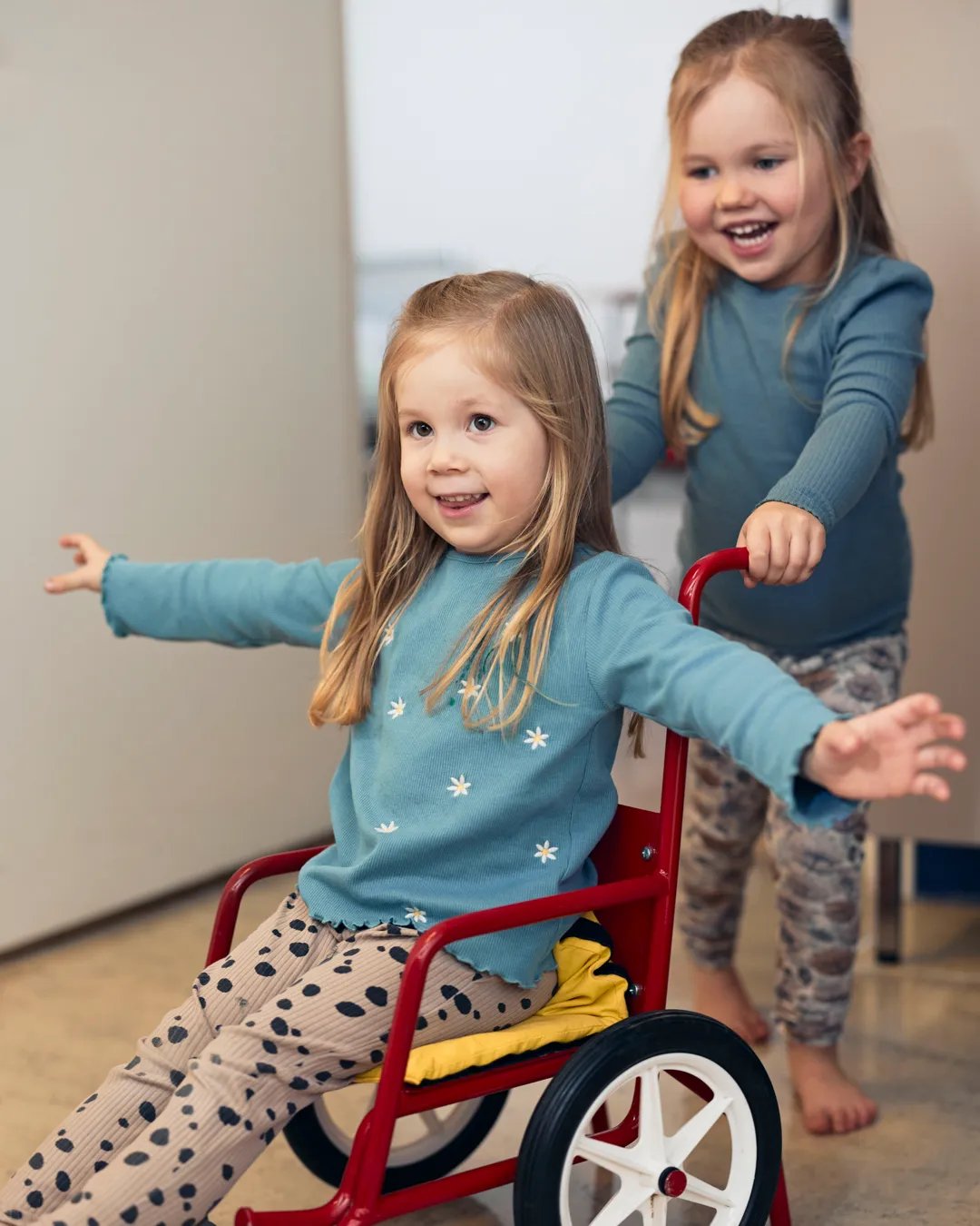 Två små flickor leker med en liten röd rullstol.